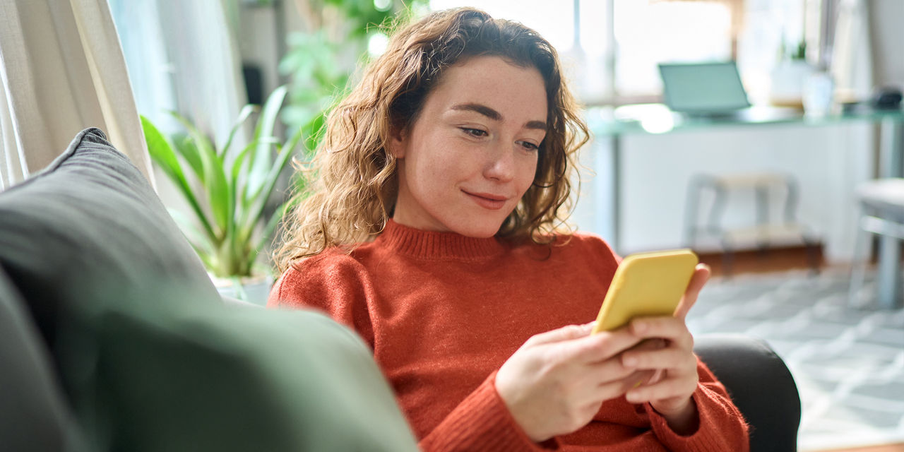 Eine junge Frau mit lockigem Haar, in einem orangefarbenen Pullover sitzt auf einem Sofa und benutzt ein gelbes Smartphone