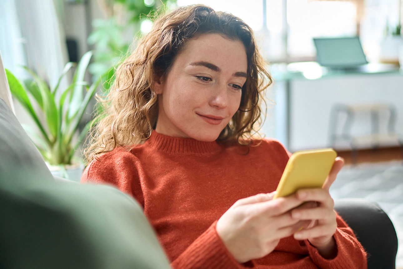 Eine junge Frau mit lockigem Haar, in einem orangefarbenen Pullover sitzt auf einem Sofa und benutzt ein gelbes Smartphone  7