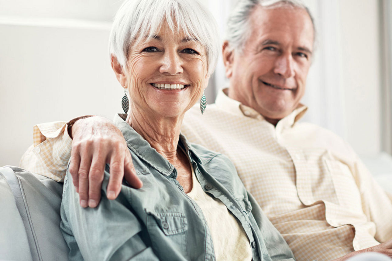 Ein glückliches älteres Paar auf einem Sofa; der Mann hat den Arm um die Frau gelegt, beide lächeln.