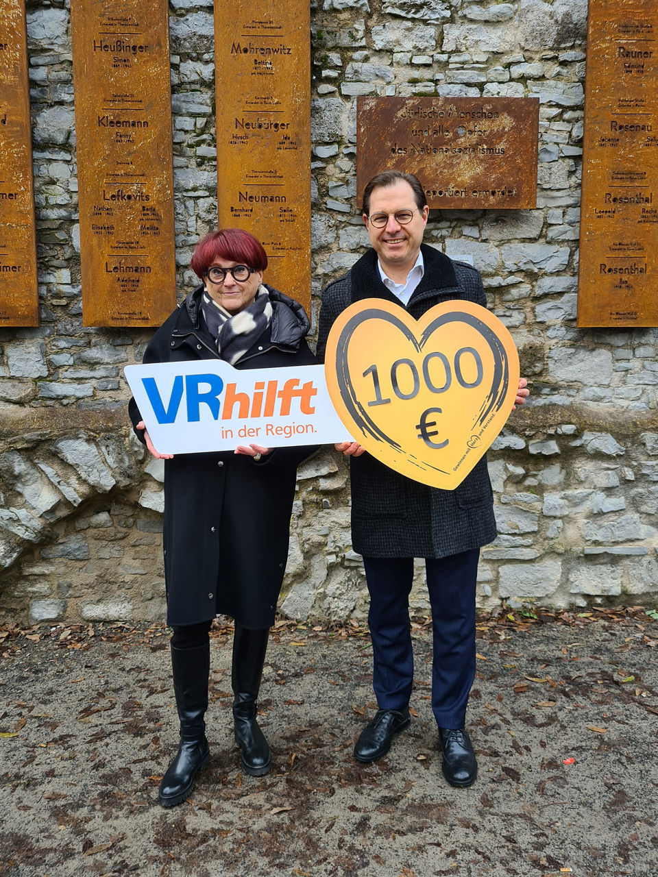 Es sind weibliche und männliche Person zu sehen. Sie halten ein großes, herzförmiges Schild mit der Aufschrift 1.000 €.