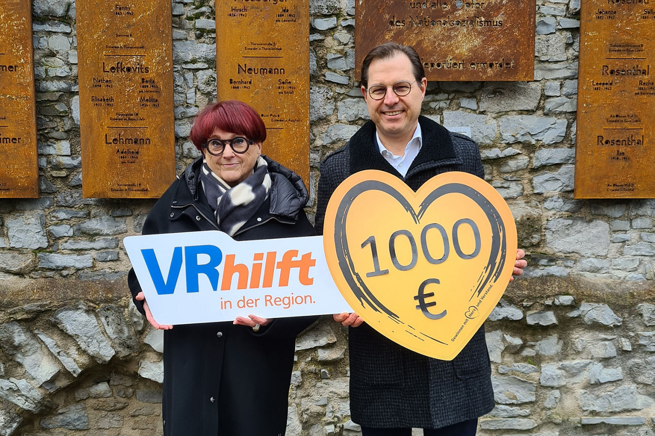 Es sind weibliche und männliche Person zu sehen. Sie halten ein großes, herzförmiges Schild mit der Aufschrift 1.000 €.