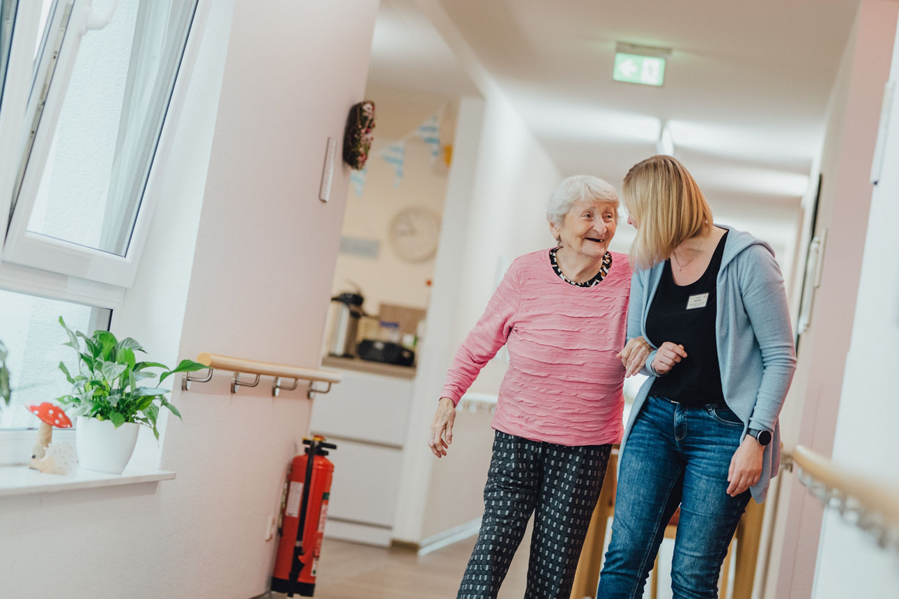 Eine Krankenschwester spaziert mit einer älteren Dame durch den Flur