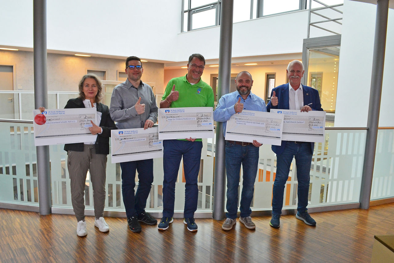 Fünf Personen halten übergroße Spendenschecks in den Händen und zeigen Daumen hoch – ein Moment der Freude und Unterstützung für gemeinnützige Projekte.
