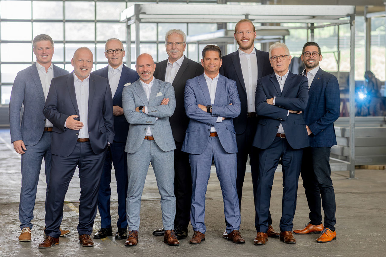 Neun Männer in Anzügen posieren lächelnd in einer Industriehalle.