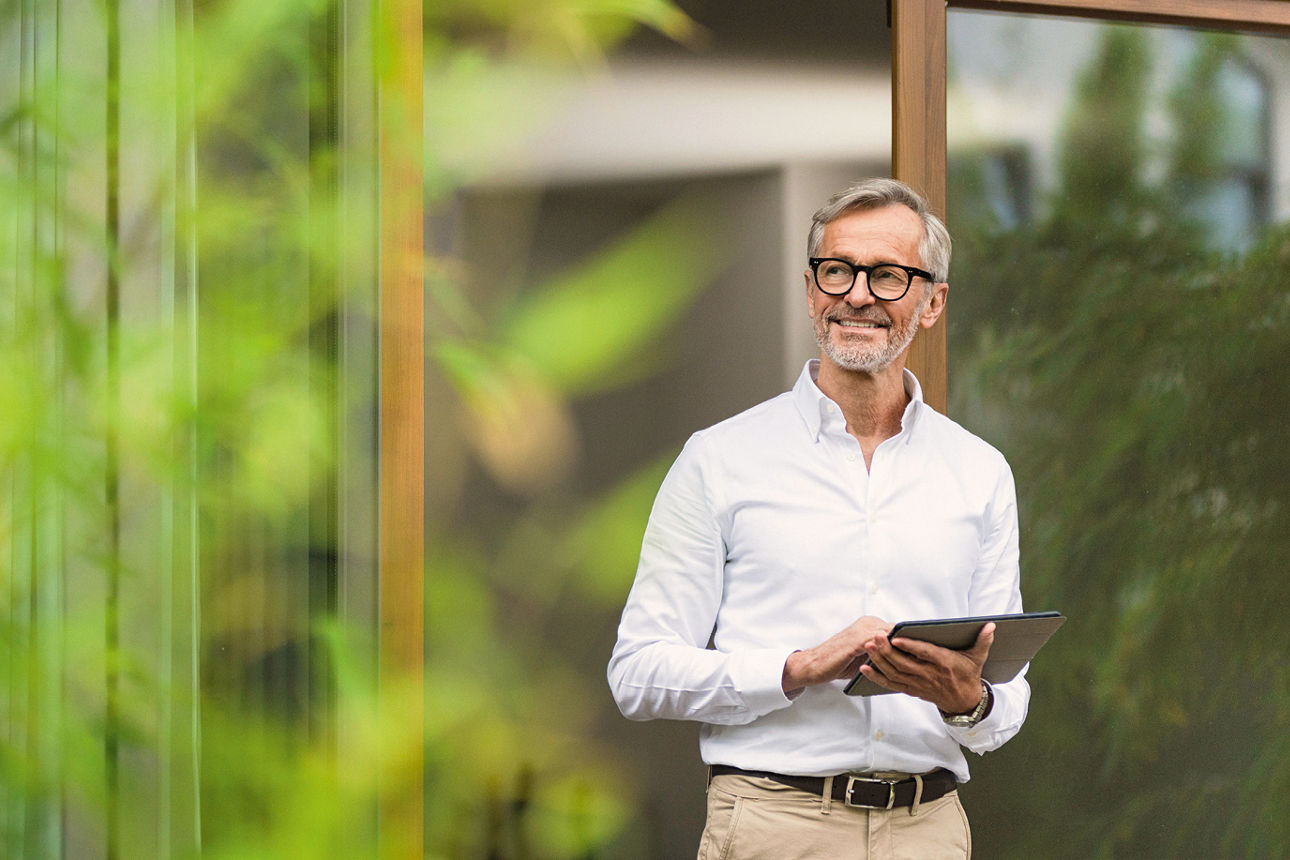Mann steht mit Tablet im Garten vor einem Haus