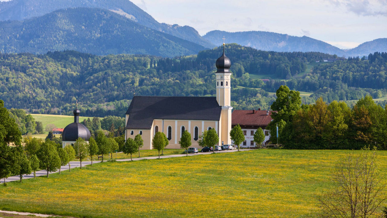 Eine malerische Landschaftsaufnahme mit einer großen, hellgelben Kirche mit dunklem Zwiebelturm in der Mitte. Sie ist umgeben von grünen Wiesen mit gelben Blumen und einer Baumallee. Im Hintergrund erheben sich bewaldete Berge unter einem teilweise bewölkten Himmel.