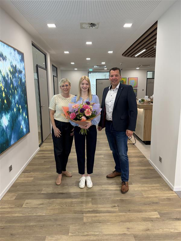 Zwei Frauen und ein Mann haben sich zum Foto aufgestellt. Die Frau in der Mitte hat einen Blumenstrauß in der Hand.