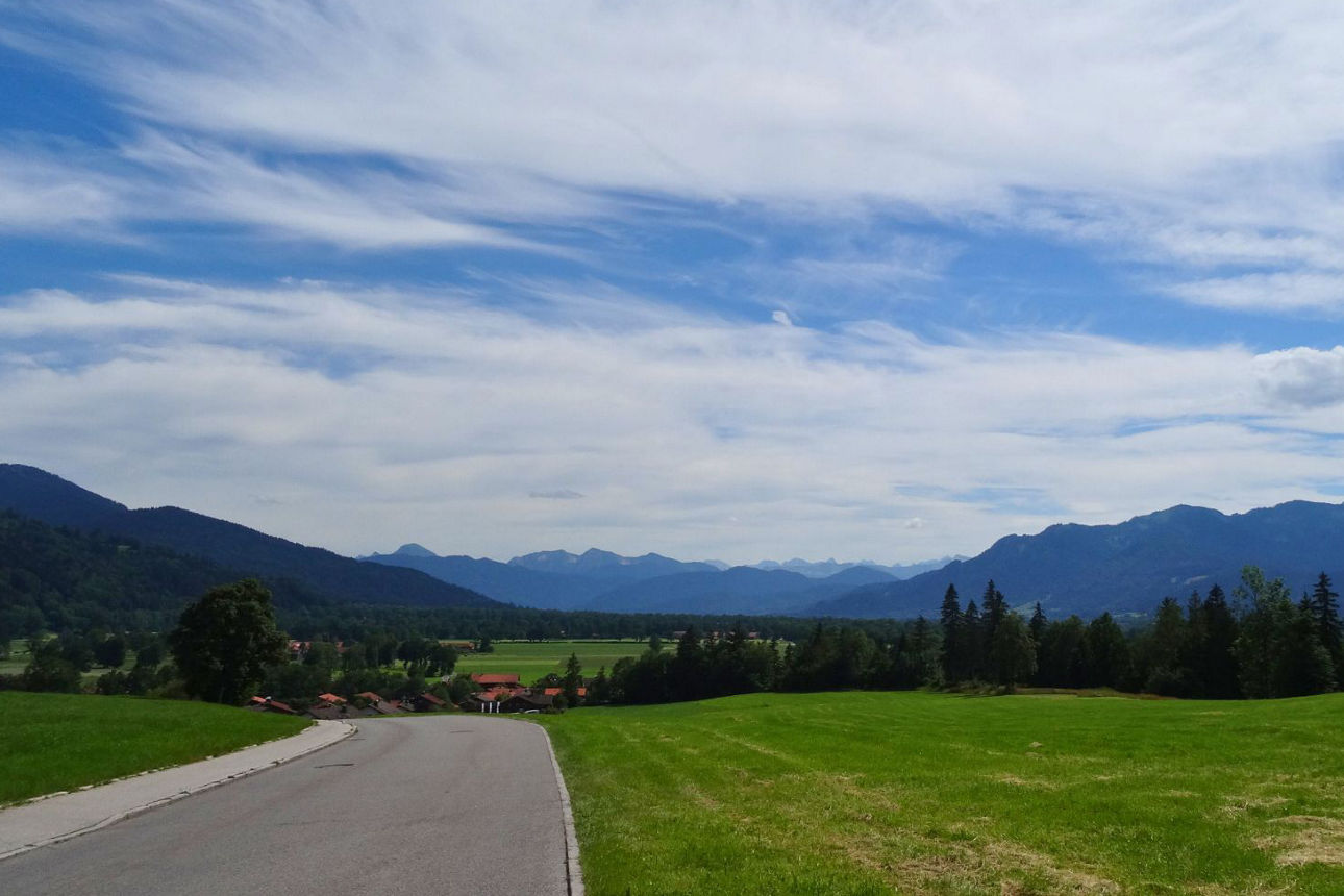 Eine weite Landschaftsansicht des Isarwinkels mit einem gewundenen Weg, der durch grüne Felder führt. Am Horizont erheben sich imposante, bewaldete Berge unter einem weiten blauen Himmel mit ausgedehnten weißen Wolken. Vereinzelt sind Häuser mit roten Dächern und Nadelbäume zu sehen.