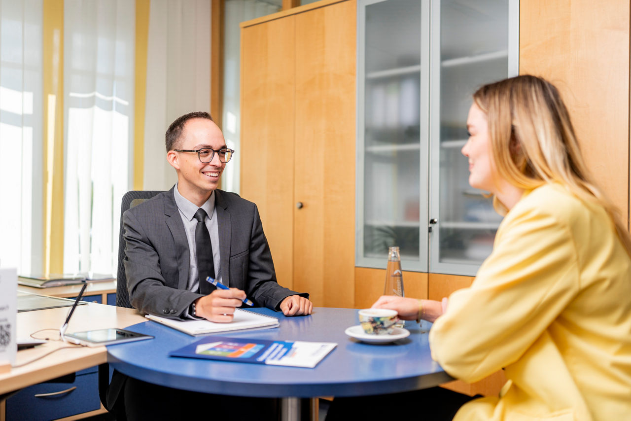 junger Herr im Anzug berät eine junge Dame mit gelben Blazer in einem Büro