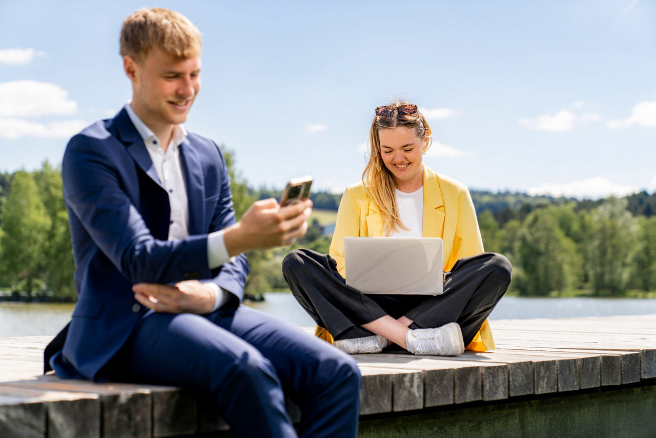 zwei junge Erwachsene sitzen mit Handy und Laptop auf einem Steg an einem See
