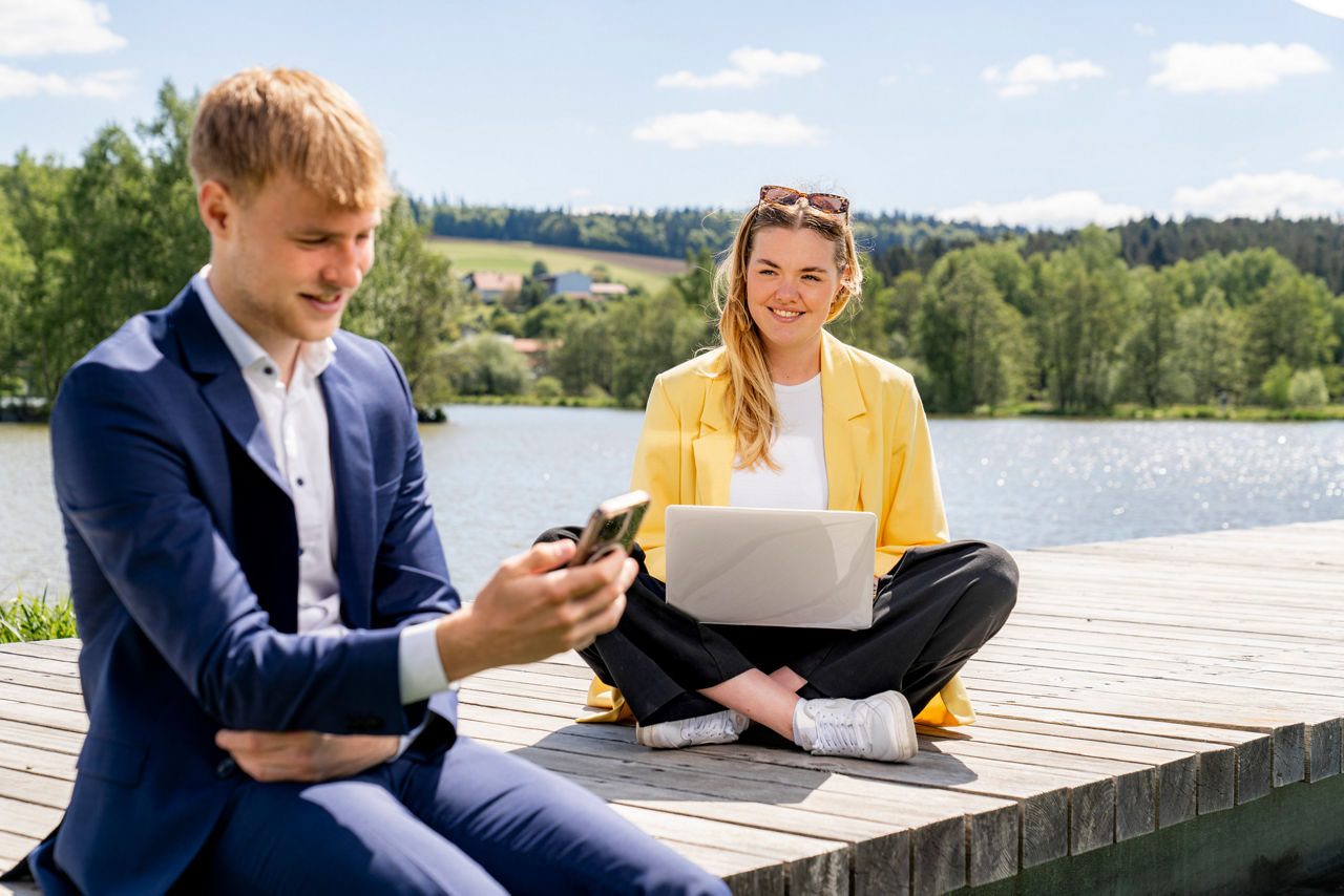 zwei junge Erwachsene sitzen mit Handy und Laptop am Steg an einem See
