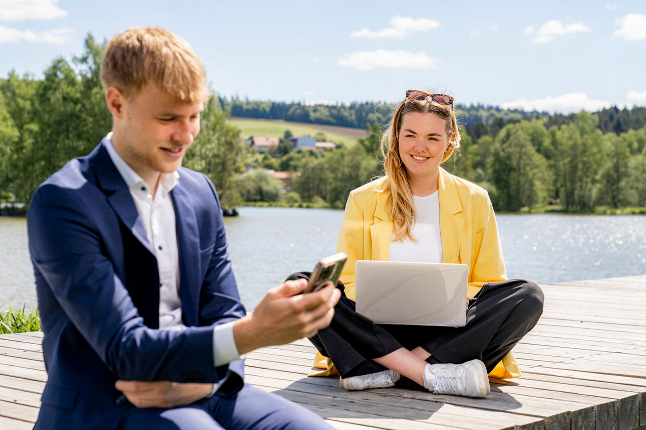 zwei junge Erwachsene sitzen mit Handy und Laptop am Steg an einem See