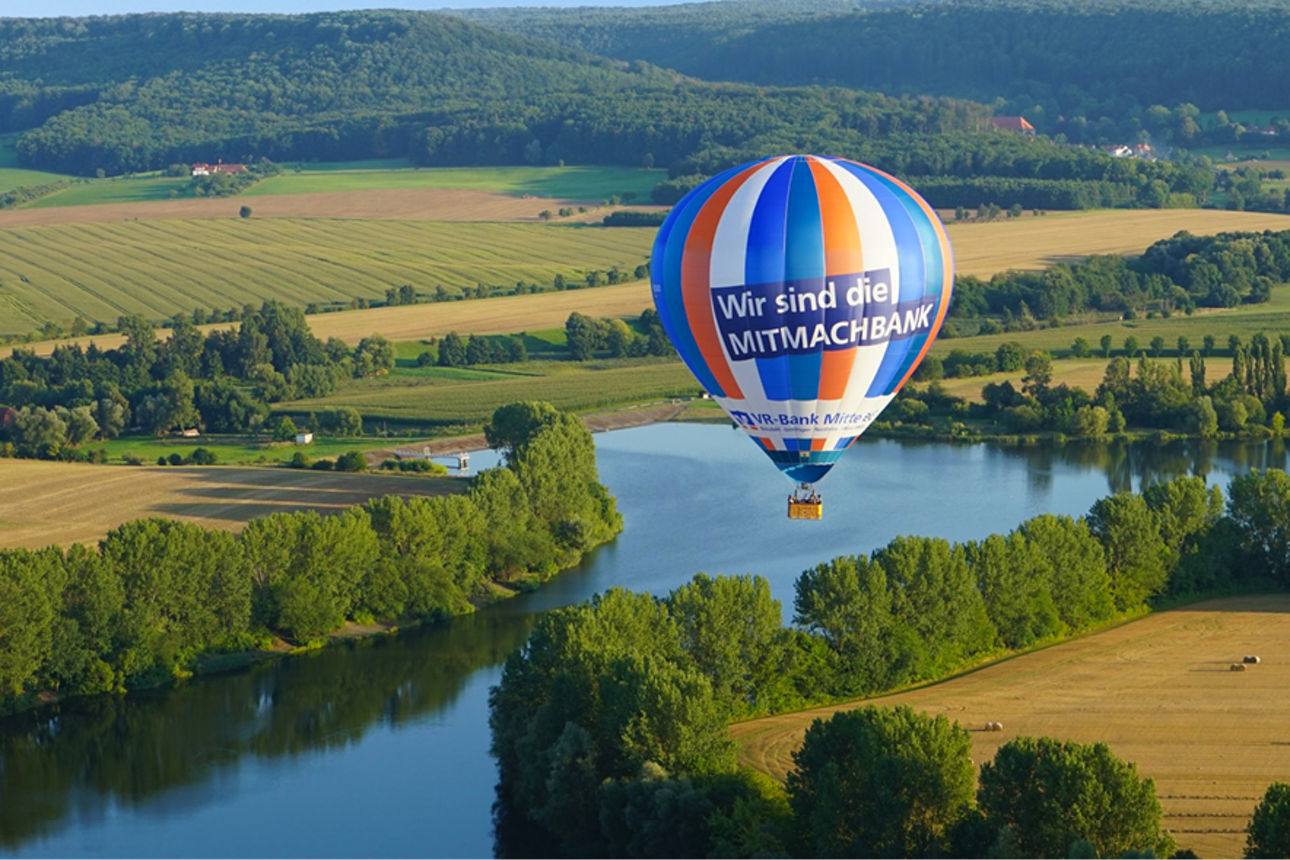 Heißluftballon fährt über einen See und mehreren Feldern um den See