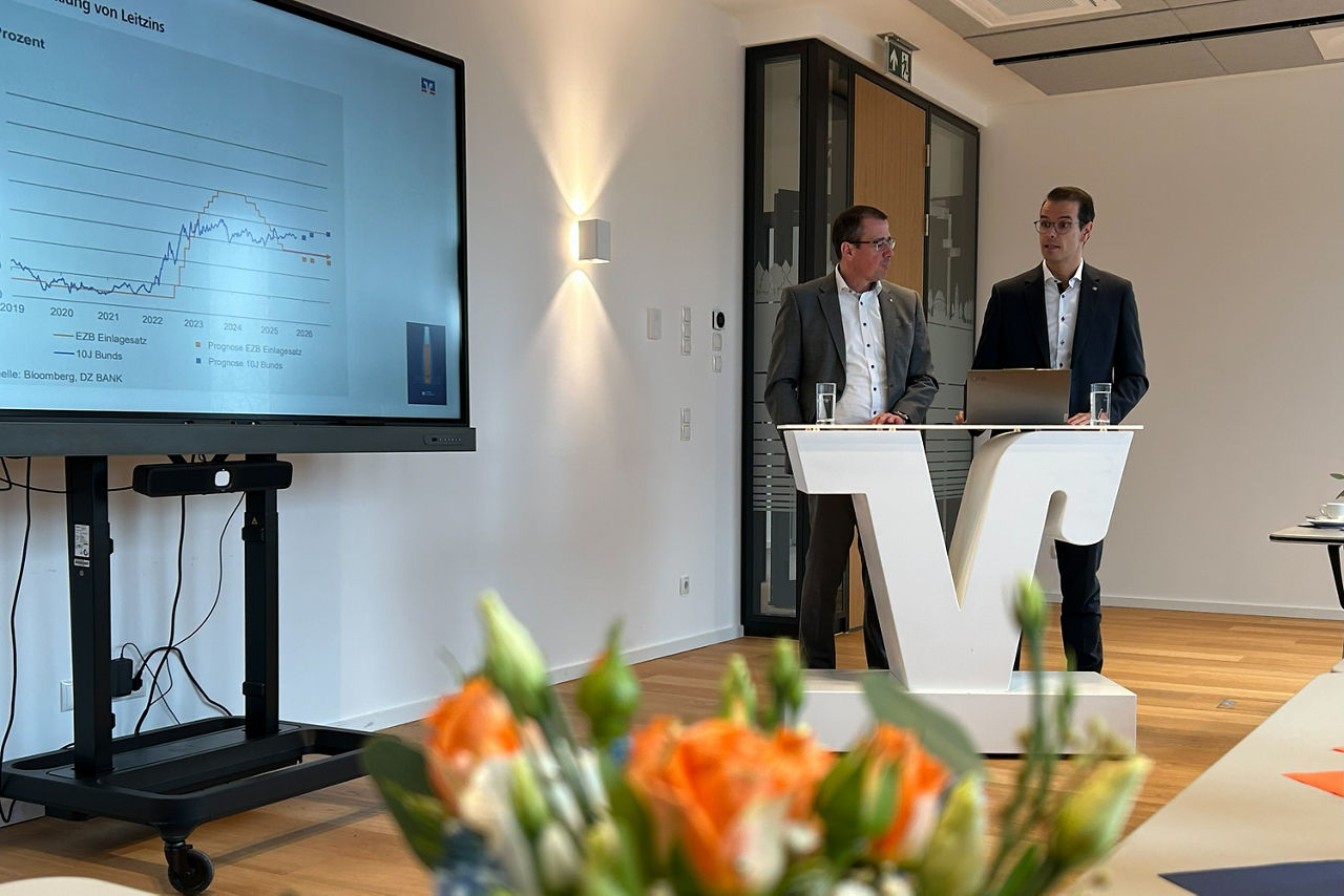 Vorstand N. Scholtholt und St. Korte stehen hinter einem Rednerpult. Links im Bild sieht man noch einen Bildschirm mit einer Präsentation.