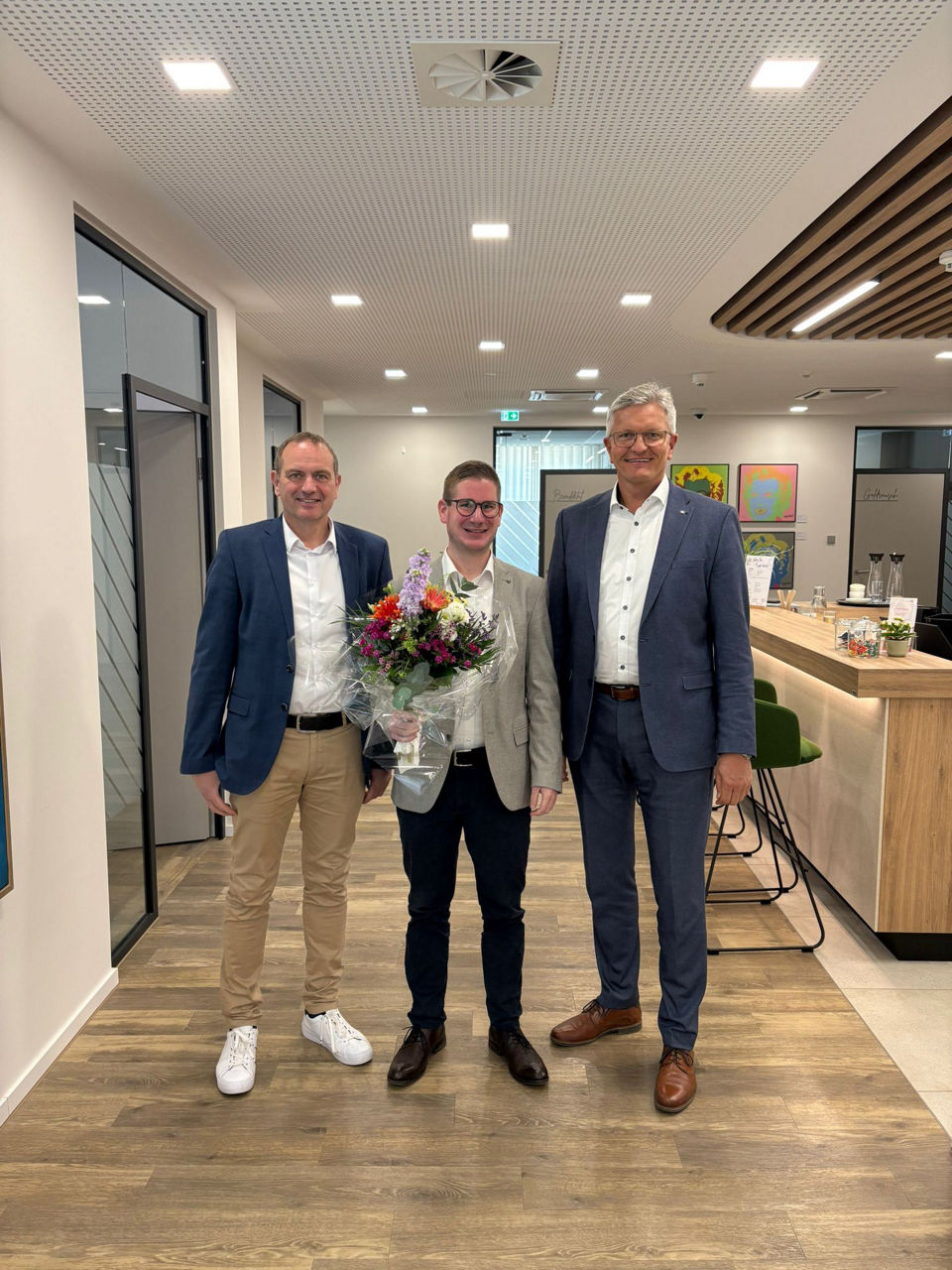 Drei Männer haben sich zum Foto aufgestellt. Der Mann in der Mitte hat einen Blumenstrauß in der Hand.