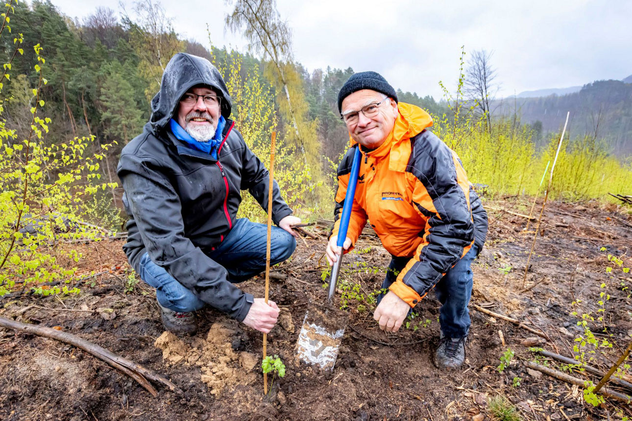 Zwei Männer in Regenkleidung hocken an einem kleinen Setzling und lächeln in die Kamera