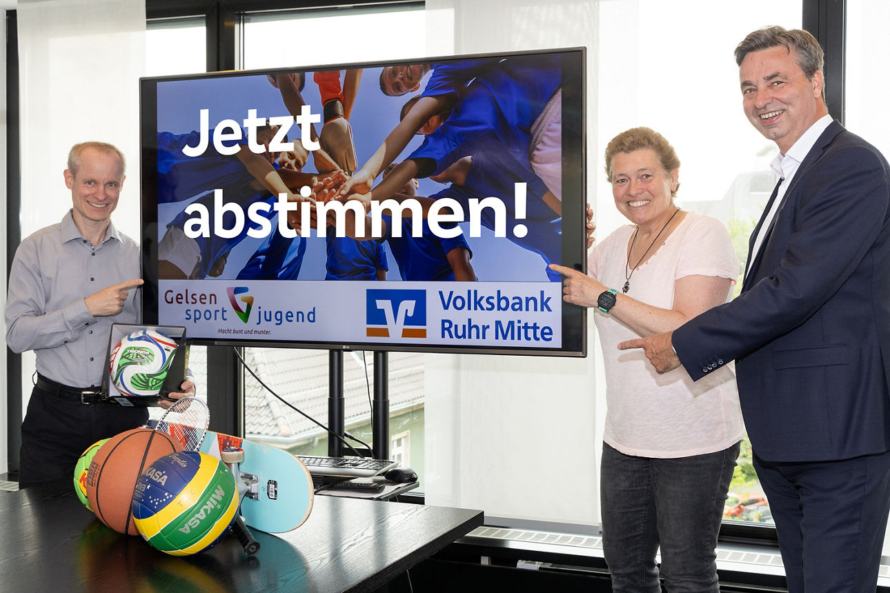 Drei Personen stehen neben einem großen Bildschirm und zeigen darauf. Auf dem Bildschirm ist der Text „Jetzt abstimmen!“ sowie Logos von Gelsensport Jugend und Volksbank Ruhr Mitte zu sehen. Im Vordergrund liegen verschiedene Sportgeräte wie ein Volleyball, Basketball, Tischtennisschläger und andere Utensilien auf einem Tisch. Die Szene findet in einem hellen Innenraum mit Fenstern im Hintergrund statt.