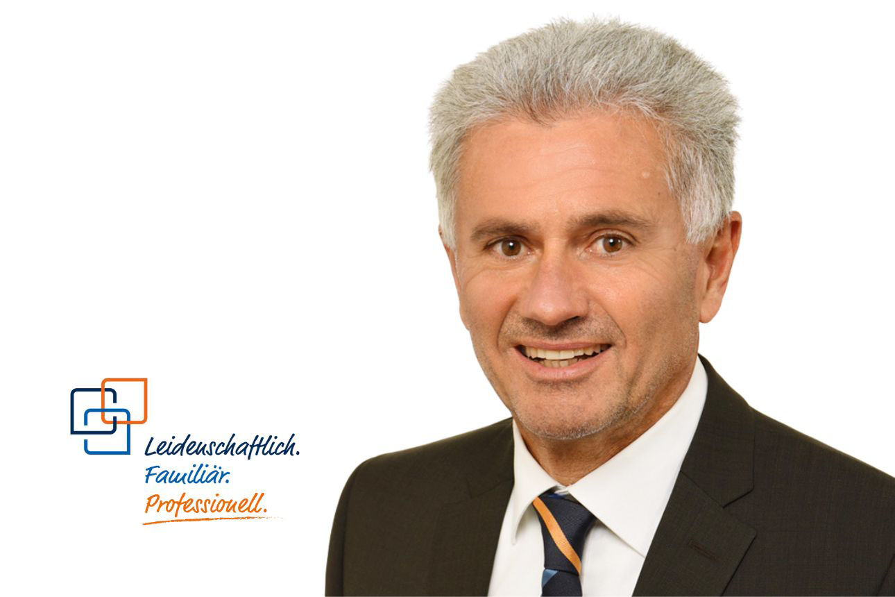 Johann Engl, ein Mann mit grau-melierten Haaren und einem freundlichen Lächeln, trägt einen dunklen Anzug mit einer gestreiften blau-orangenen Krawatte und blickt direkt in die Kamera.