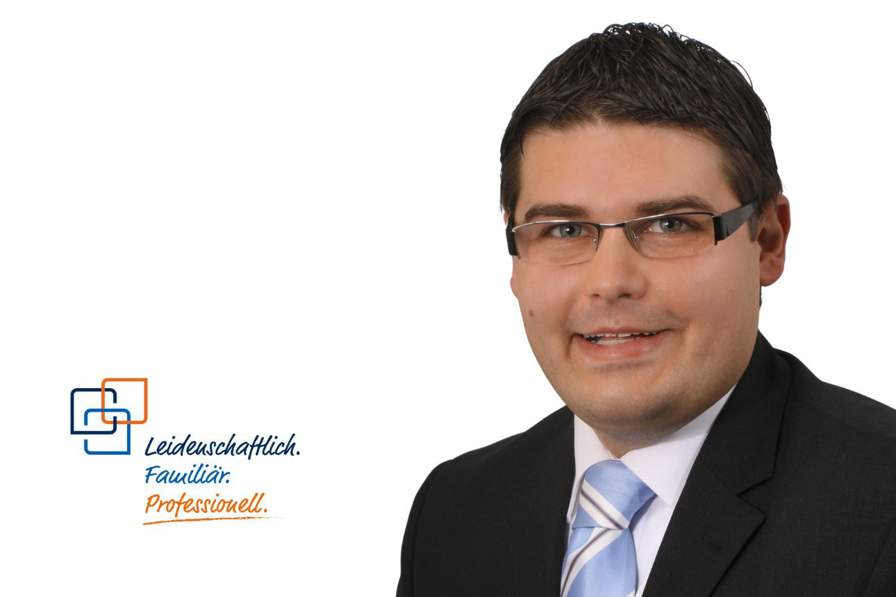 Josef Geißinger, ein Mann mit Brille und kurzen, dunklen Haaren, lächelt und trägt einen dunklen Anzug mit einer gestreiften blau-weißen Krawatte.