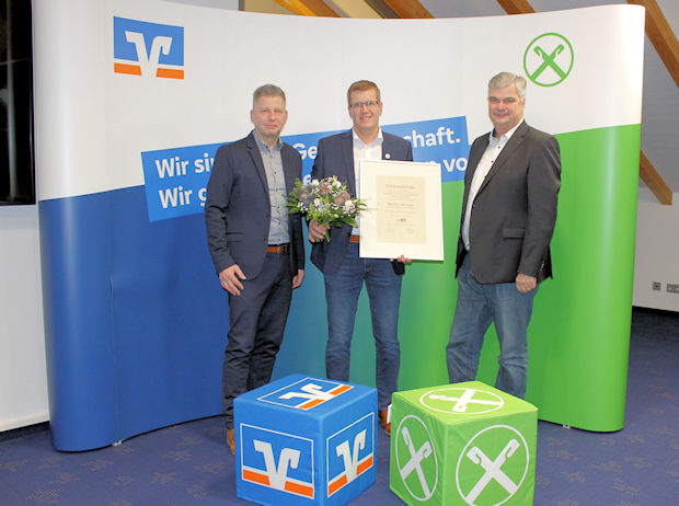 Drei Männer stehen vor einem blau-grünen Hintergrund und halten eine gerahmte Urkunde und einen Blumenstrauß in der Hand.
