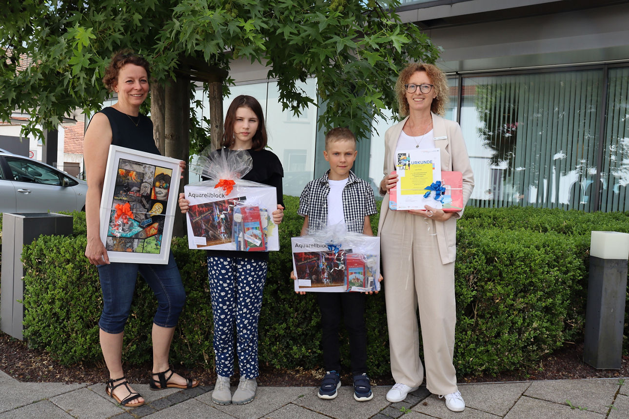 Zwei Erwachsene und zwei Kinder stehen mit Urkunden und Preisen in den Händen vor einem Baum und einem modernen Gebäude. Es handelt sich um die Ehrung der Gewinnerinnen und Gewinner des Jugendwettbewerbs ‚jugend creativ‘.