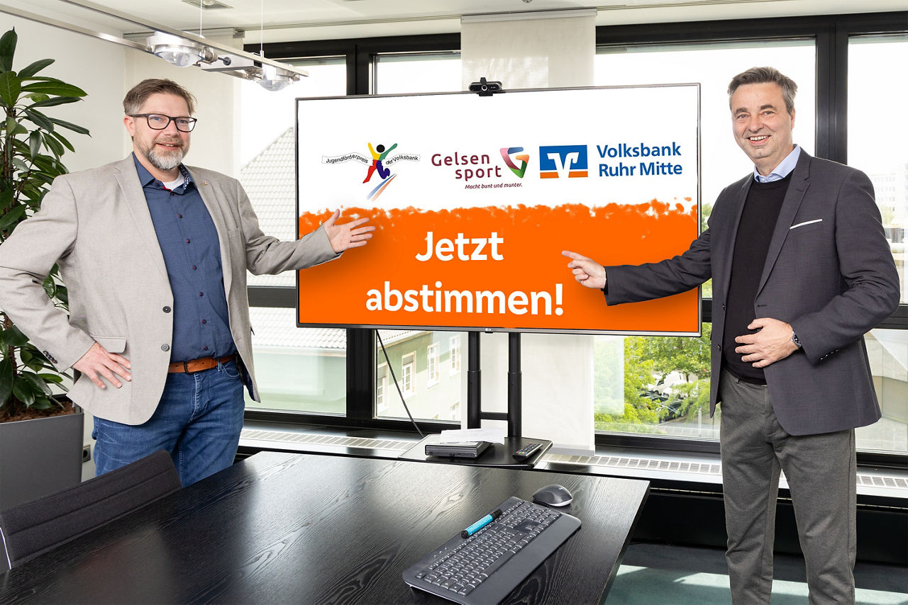 Zwei Männer im legeren Anzug lächeln für ein Pressefoto zum Jugendförderpreis der Volksbank Ruhr Mitte für Gelsenkirchen in die Kamera. Links steht Marc Kopatz von Gelsensport und zeigt auf den Fernseher zwischen den beiden Personen. Rechts steht Jörg Lott, Vorstand der Volksbank und zeigt ebenfalls auf den Fernseher. Auf dem Fernseher sind die Logos vom Jugendförderpreis, Gelsensport und der Volksbank zu sehen. 