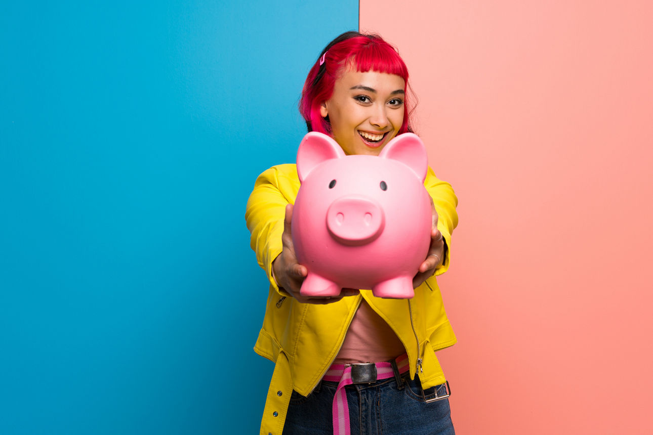 Junge Frau steht vor einer blau-rosa Wand und hält ein großes rosanes Sparschwein hoch.