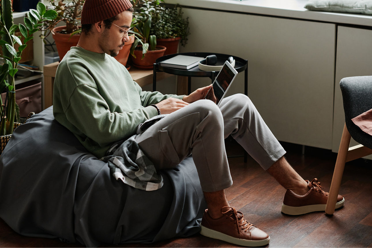 Ein junger Mann sitzt auf einem Sitzsack in seiner Wohnung und hat ein Tablet in der Hand.
