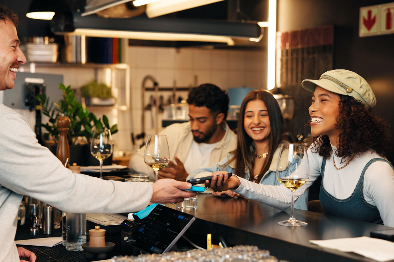 Mehrere Personen sitzen an einer Bar sitzen, während eine Person eine kontaktlose Zahlung durchführt.