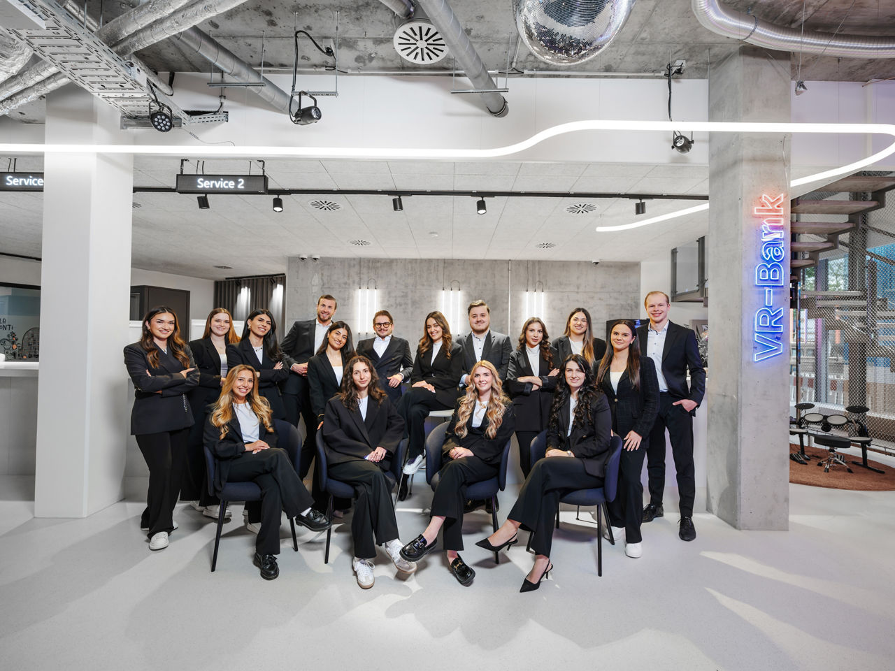 Gruppenfoto junger Bankmitarbeitender in modernem Büro mit industriellem Design und VR-Bank-Schriftzug.