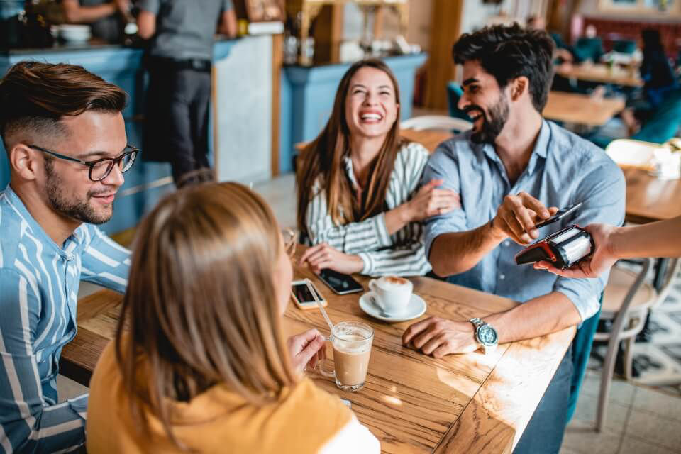 Eine Gruppe von vier jungen fröhlichen Erwachsenen sitzt in einem Café. Einer davon bezahlt mit seinem Smartphone