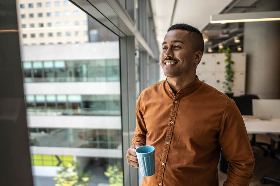 Junger Mann steht in enem Büro am Fesnter mit einer Tasse in der Hand