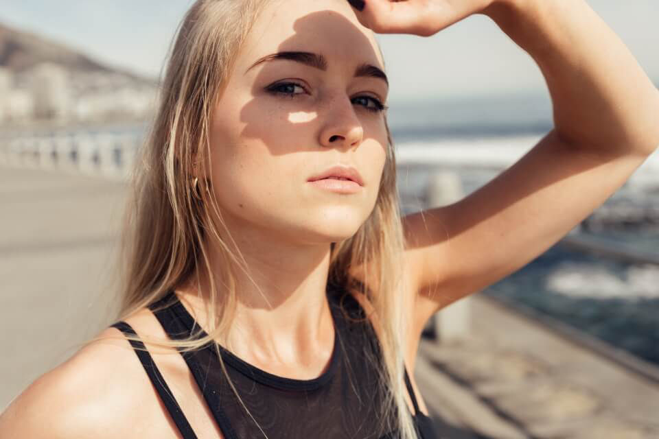 Junges Frau steht an einem Strand und schützt sich mit der Hand vor der Sonne.