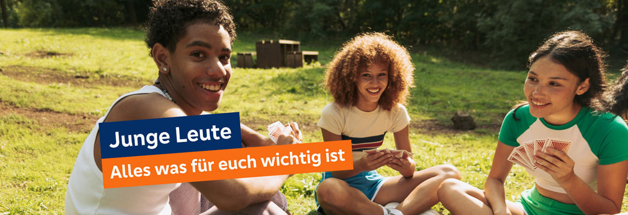 Drei junge Leute sitzen im Gras in einem Park und spielen Karten.
