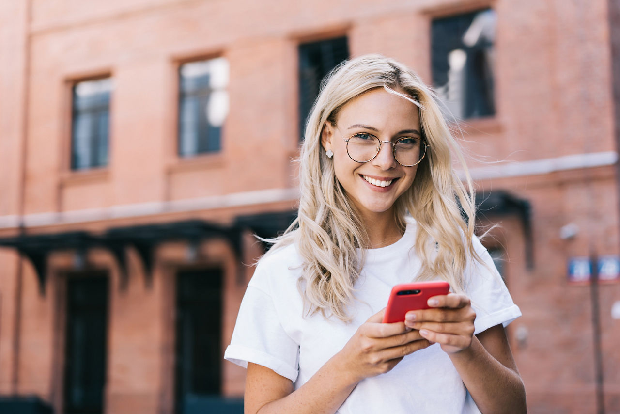 Junge Dame mit Brille lächelt in die Kamera und surft auf dem Mobiltelefon, während sie inder Stadt ist