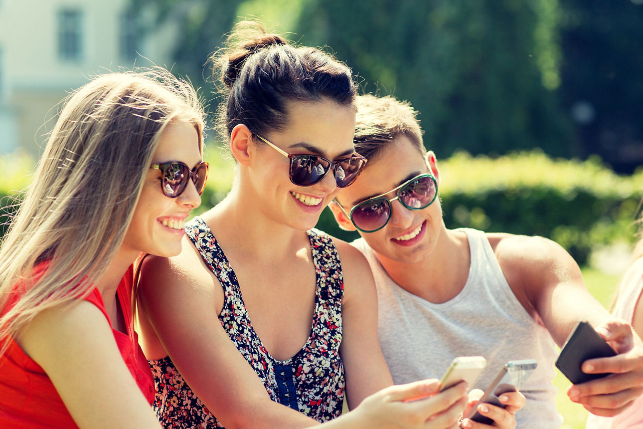 Drei Freunde mit Sonnenbrillen sitzen im Freien und schauen gemeinsam auf ihre Smartphones.