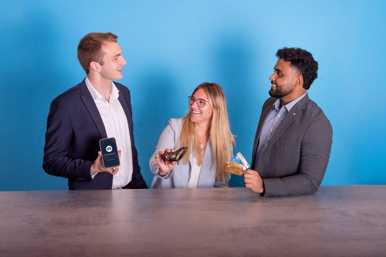 Drei junge Leute stehen nebeneinander. Jeder hält etwas anderes in der Hand: - ein Smartphone - einen Bronzestier - Kreditkarten
