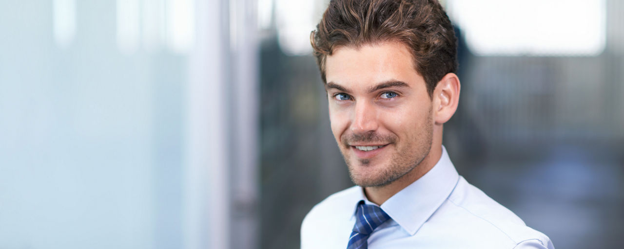 Ein junger Mann steht in einem hellen Büroflur und trägt Hemd und Krawatte.