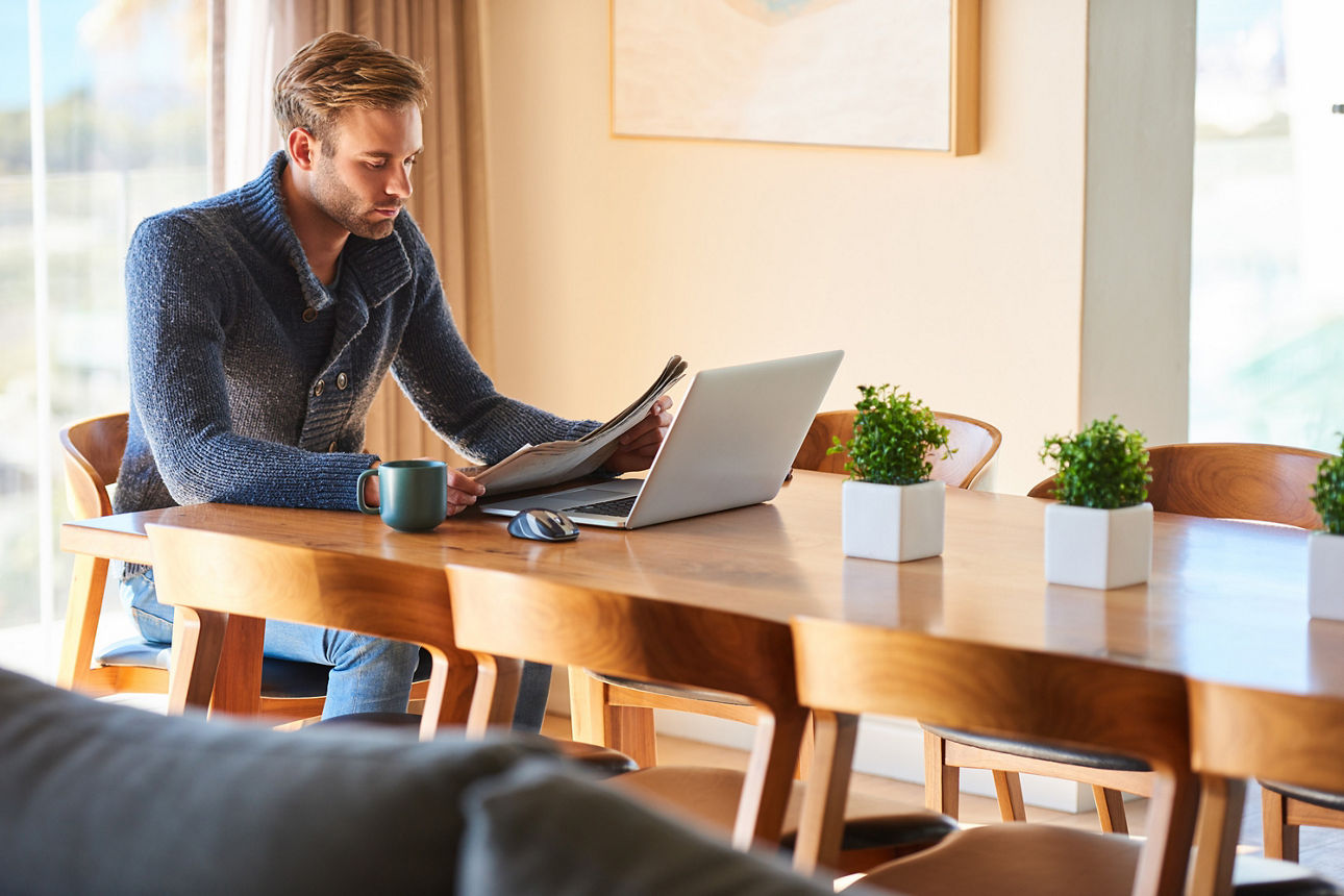 Junger Mann sitzt morgens mit einer Tasse Kaffee an seinem Frühstückstisch und liest die Zeitung. Neben ihm steht sein Laptop