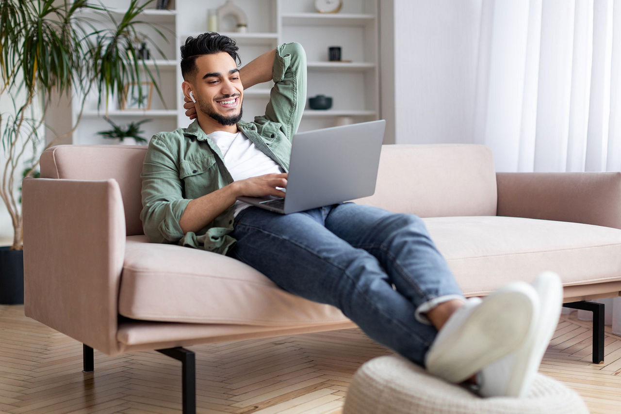 Junger Mann mit Bart sitzt mit Laptop auf dem Sofa
