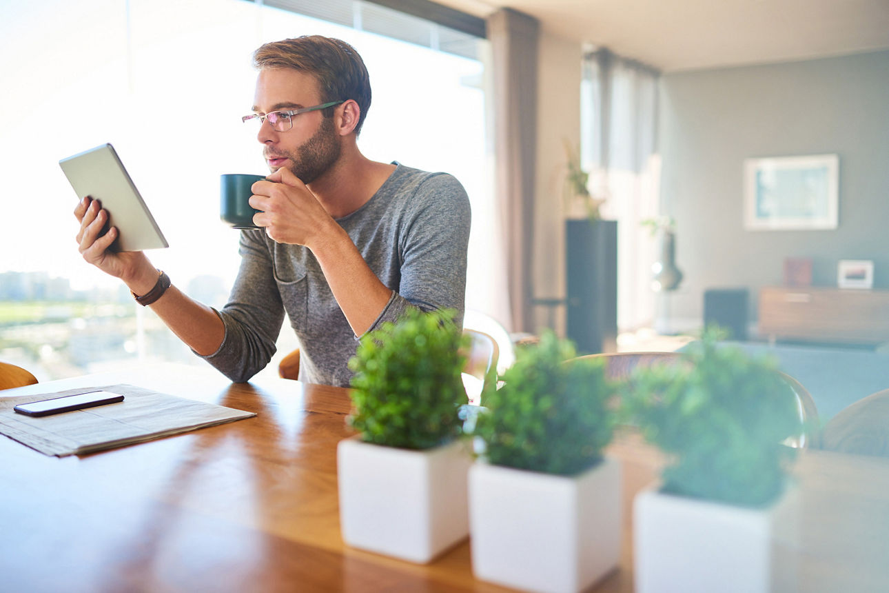 Junger Mann sitzt an einem Tisch im Wohnzimmer und trinkt Kaffee, während er auf einem Tablet liest. 