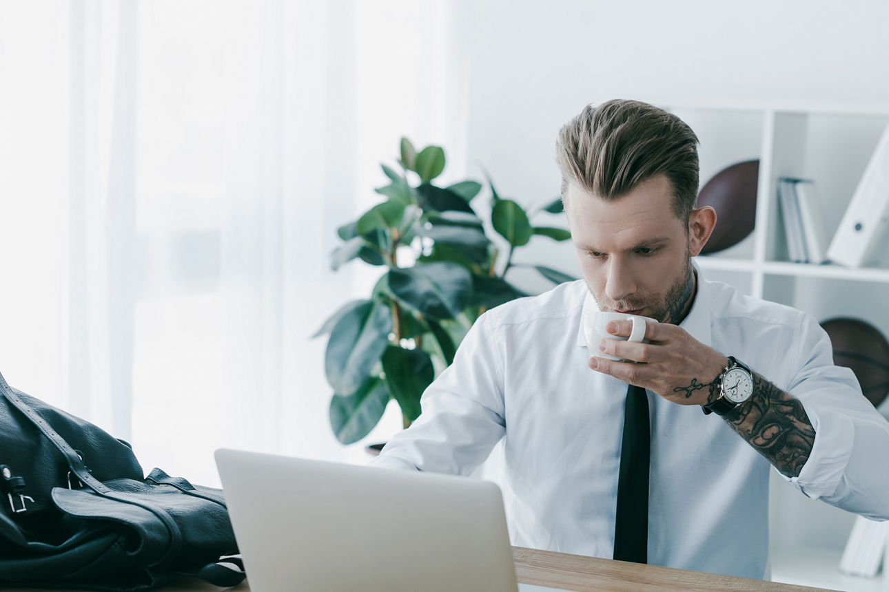 Junger Mann im Büro sitzt vor dem Laptop und trinkt eine Tasse Kaffee