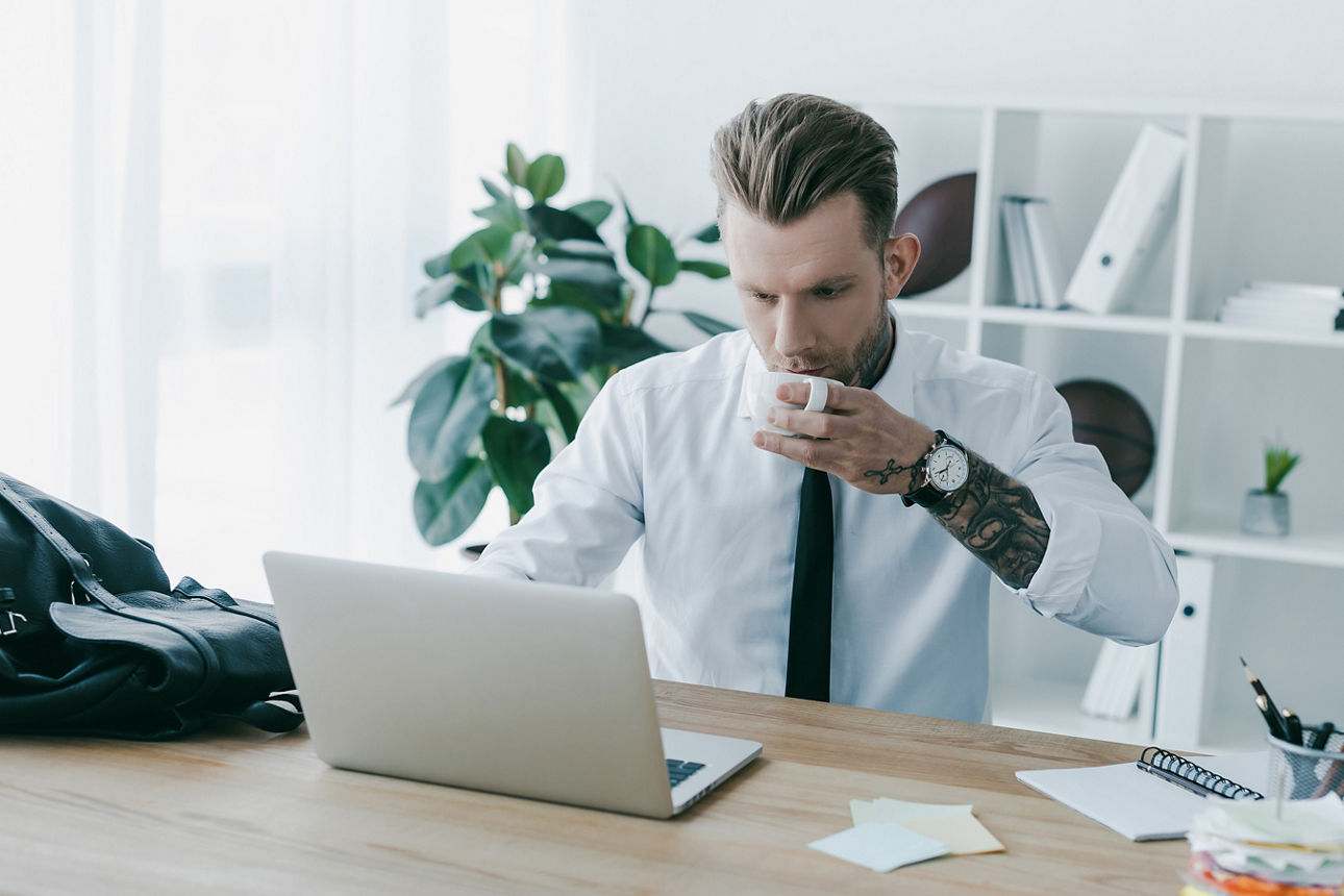 Junger Mann im Büro sitzt vor dem Laptop und trinkt eine Tasse Kaffee