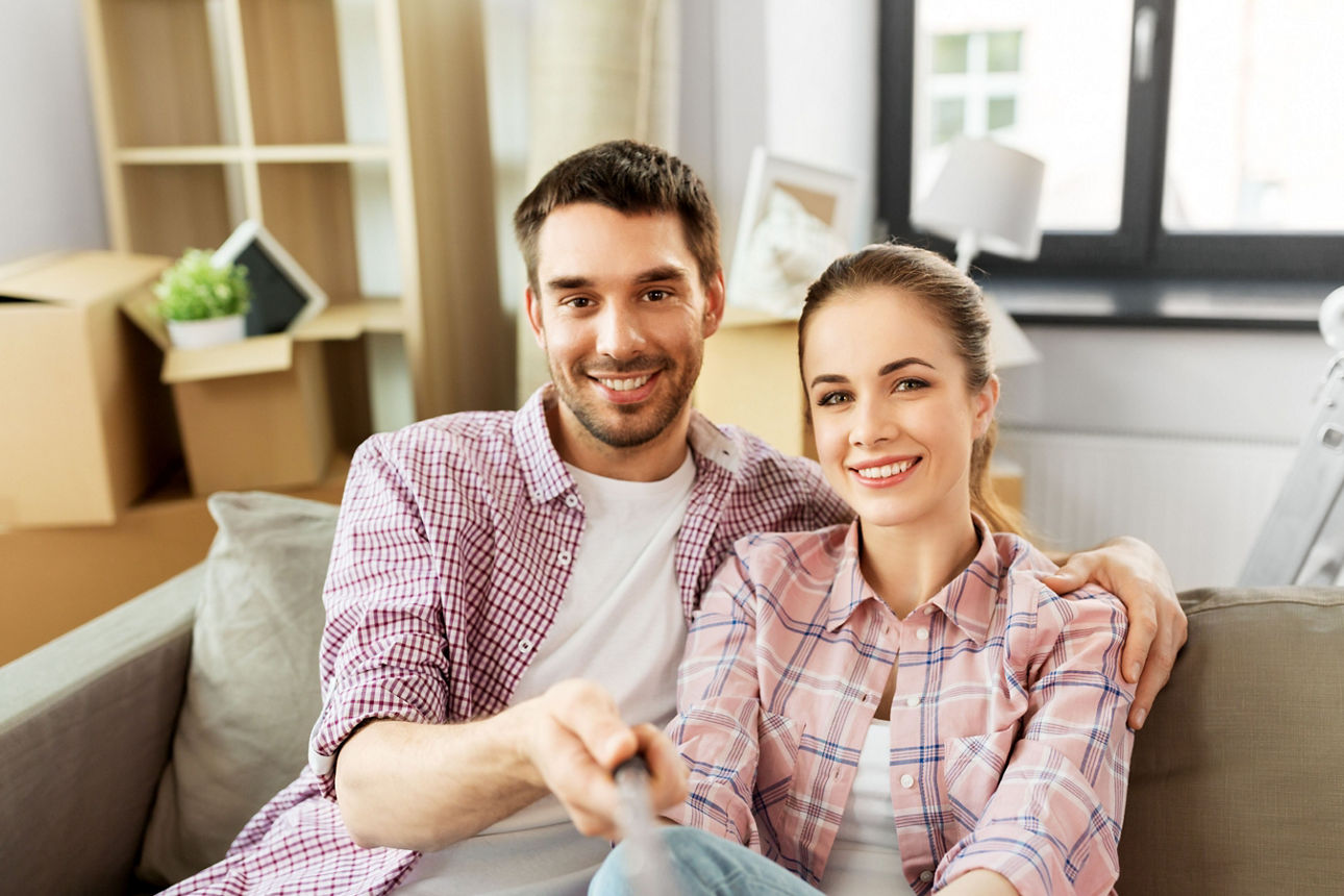 Junges Paar sitzt auf dem Sofa und lächelt im Hintergrund Umzugskartons