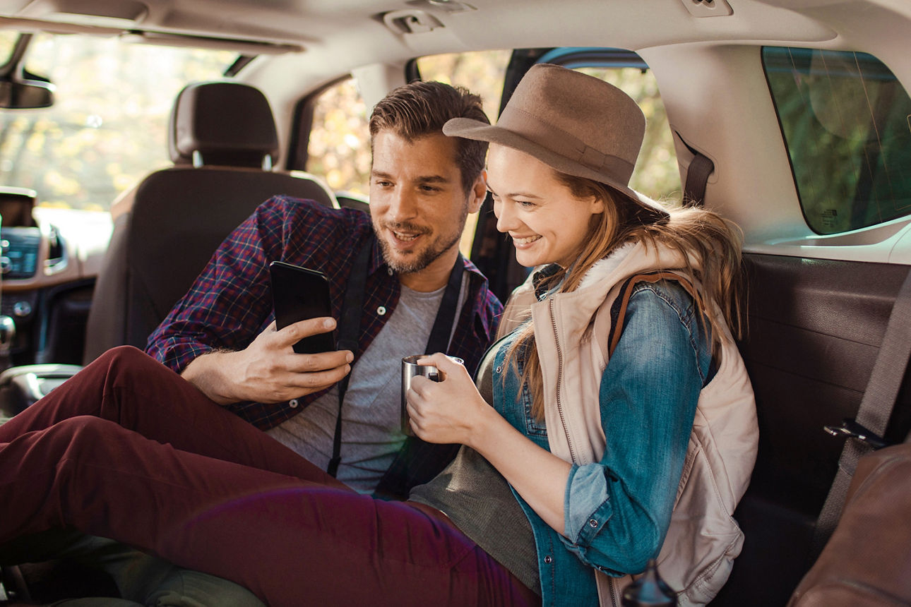 Junges Paar sitzt auf der Rückbank seines Autos und schaut gemeinsam in ein Handy