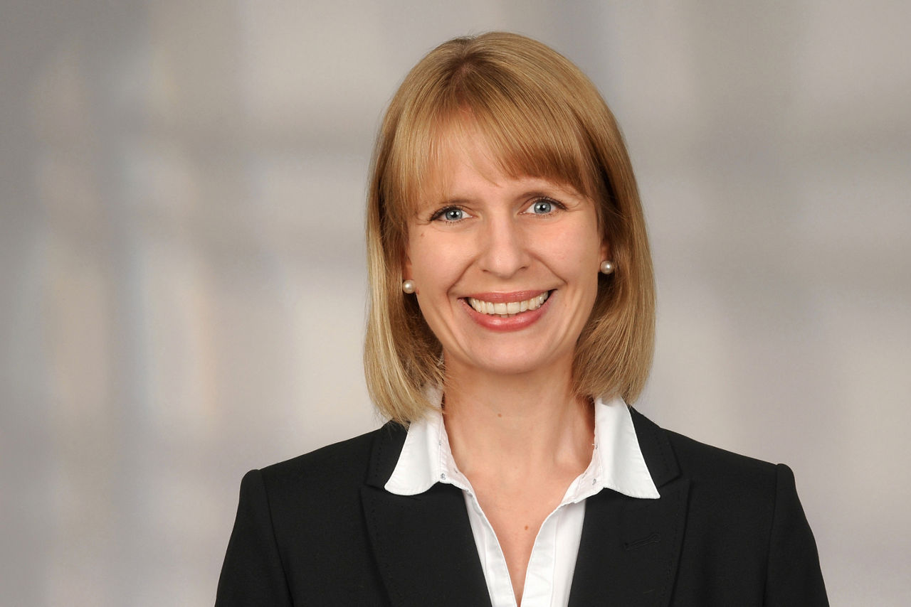 Frau mit schulterlangem blonden Haar trägt schwarzen Blazer und weiße Bluse vor neutralem Hintergrund