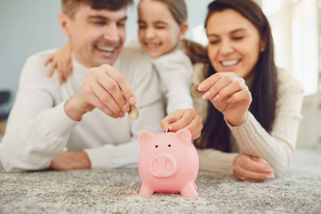 Familie legt gemeinsam Münzen in ein rosa Sparschwein auf einem Tisch und lacht dabei.