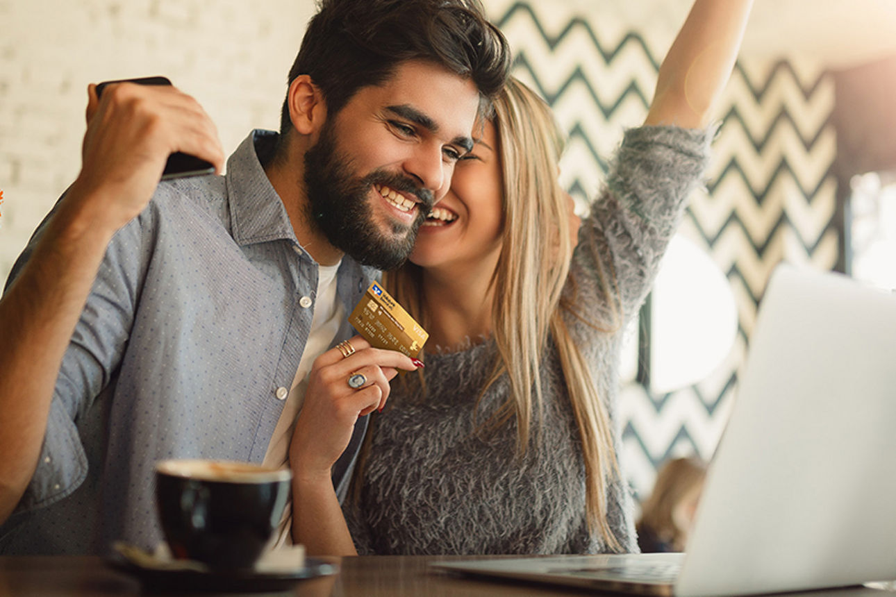 Zwei Junge Menschen sitzen jubelnd mit einer Kreditkarte vor einem Laptop.