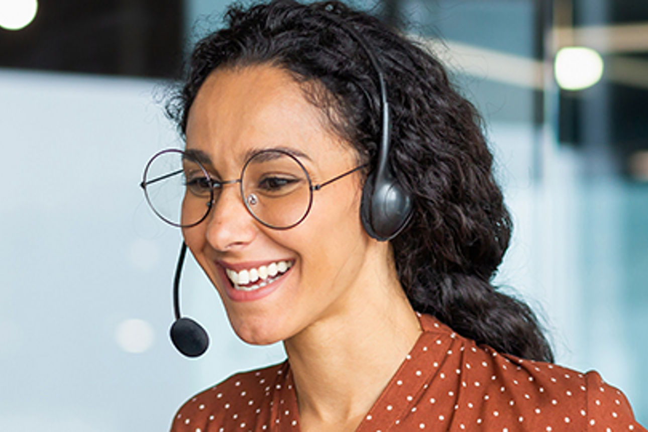 Eine Frau arbeitet in einem modernen Büro, lächelt und spricht remote über ihren Laptop und ein Headset