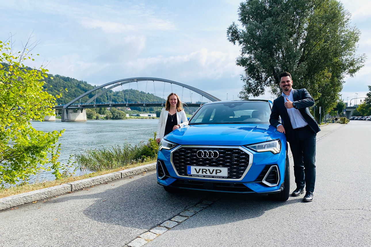Zwei Personen stehen lächelnd neben einem blauen Audi an einem Fluss mit Brücke im Hintergrund